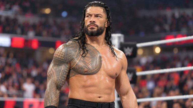Roman Reigns in black ring gear beside the ropes in a wrestling arena.
