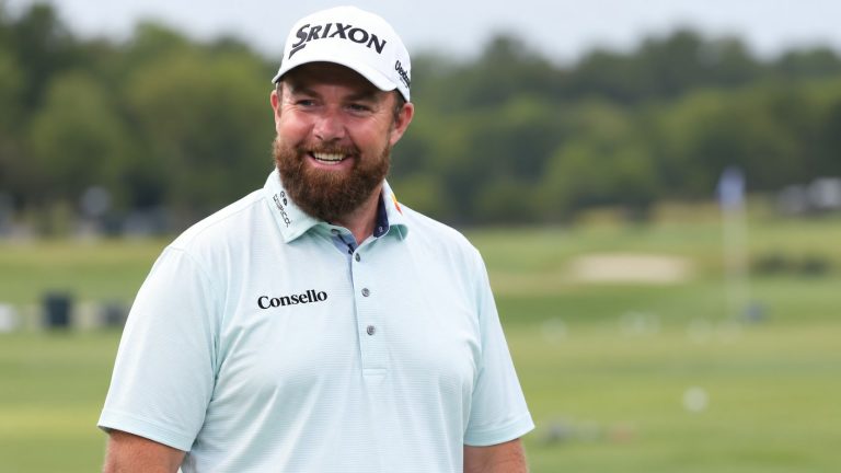 Shane Lowry wearing a white Srixon cap and pale blue golf polo beside a practice ground.