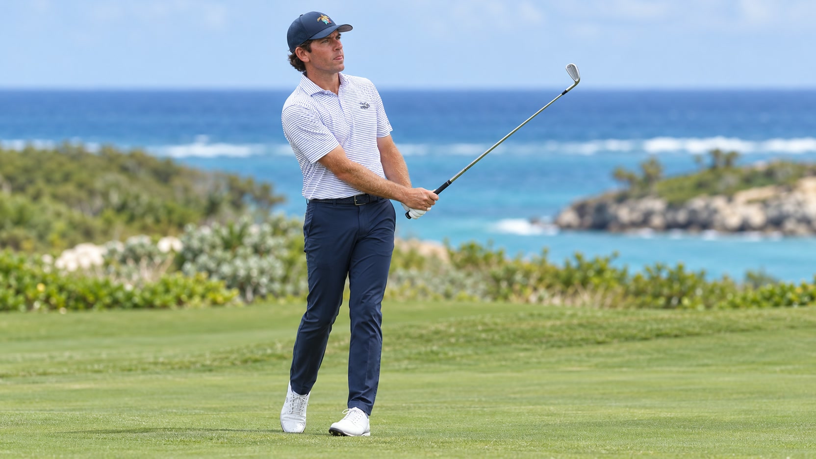 Ben Kohles in a navy cap, striped white polo and navy trousers holding an iron on a coastal golf course.