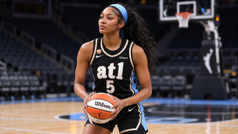 Angel Reese wearing a black Atlanta Dream No. 5 uniform and blue headband, holding a WNBA basketball on an indoor court.