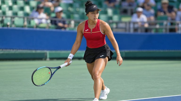 Ann Li in a red Joma top, black skirt and black visor holds a tennis racket on an outdoor hard court.