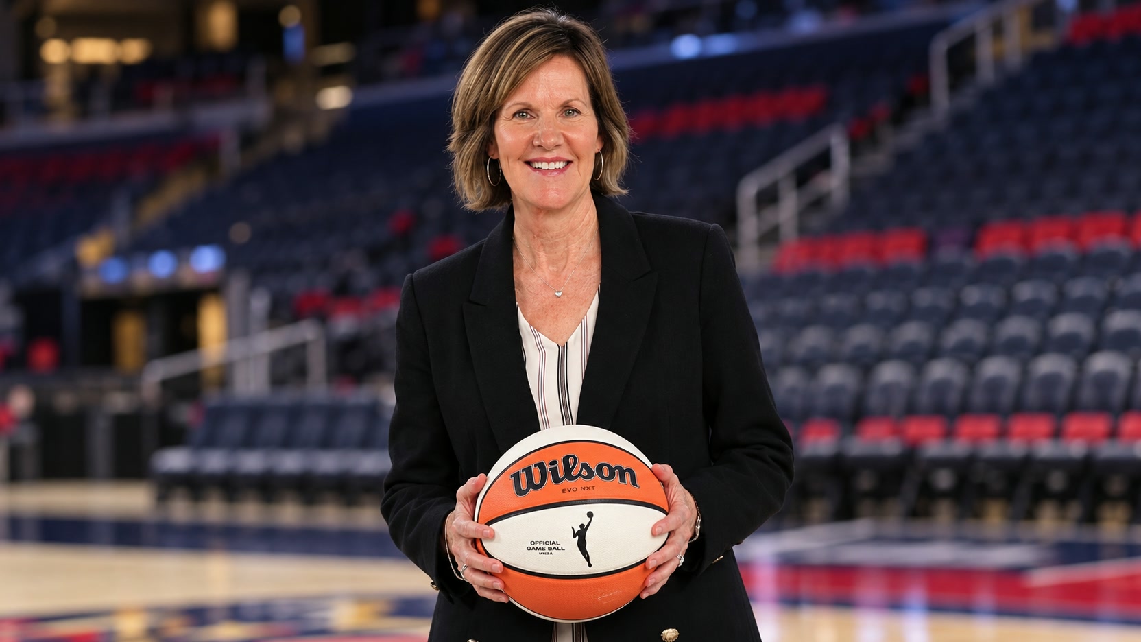 Kelly Krauskopf Kelly Krauskopf in a black blazer holding a Wilson WNBA basketball in an arena.