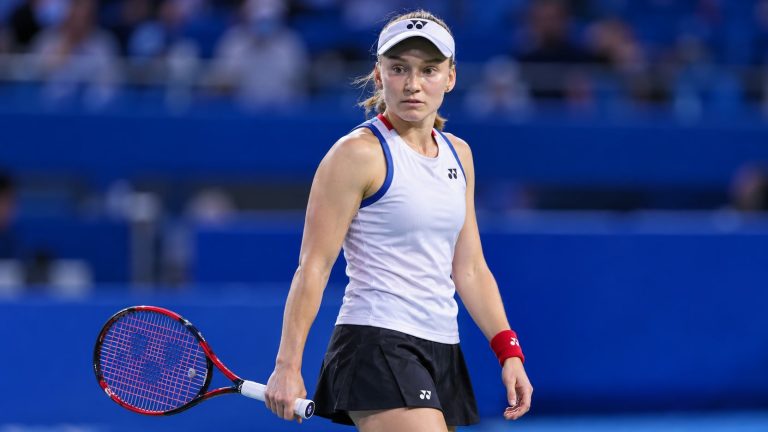 Elena Rybakina in a white Yonex visor and top holding a tennis racket on a blue hard court.