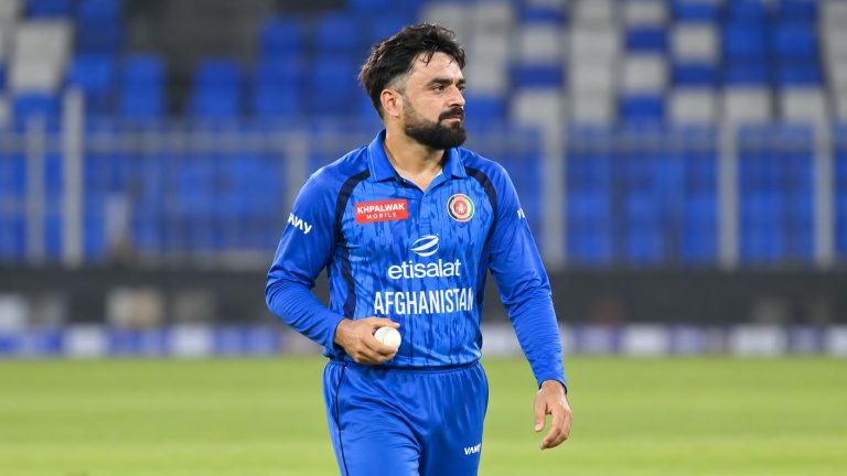 Rashid Khan in Afghanistan's blue cricket uniform holding a white ball on the field.