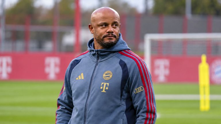 Vincent Kompany in a grey-blue Bayern Munich training jacket beside a football pitch.