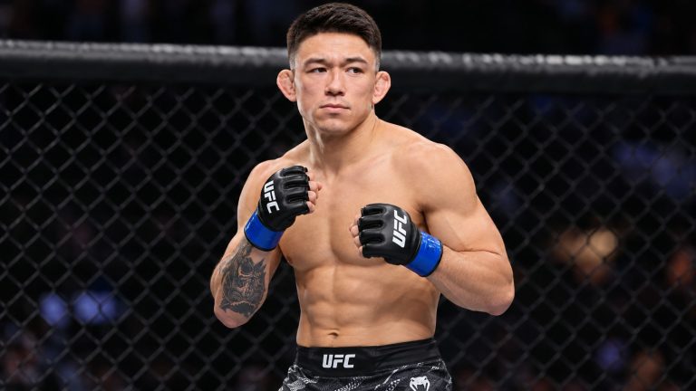 Kody Steele in black UFC gloves with blue wrist tape standing inside an MMA cage.