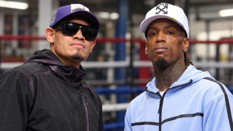 Emanuel Navarrete in a black tracksuit and purple cap beside O’Shaquie Foster in a light blue tracksuit and white cap, with boxing ring ropes behind them.