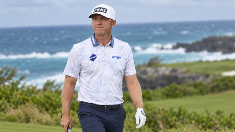 Seamus Power in a white PING cap and patterned polo holding a golf club beside a coastal fairway.
