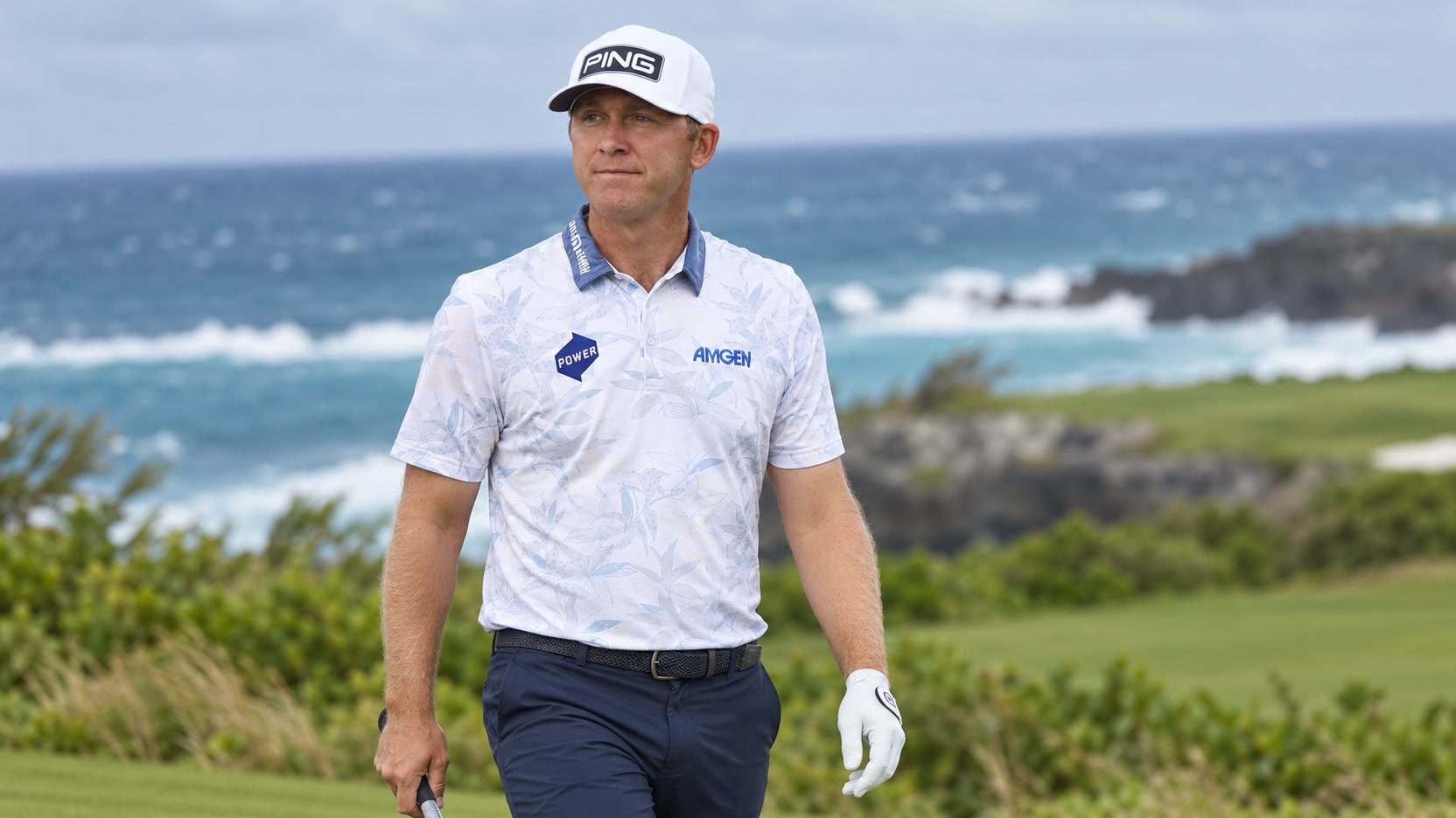 Seamus Power at a coastal golf course Seamus Power in a white PING cap and patterned polo holding a golf club beside a coastal fairway.