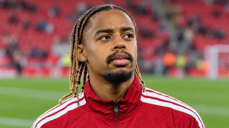 Bradley Barcola in a red Adidas training jacket with white shoulder stripes beside a football pitch.