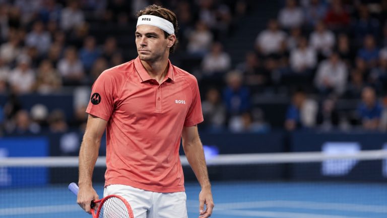 Taylor Fritz wearing a coral BOSS tennis shirt and white headband, holding a red racket on an indoor hard court.