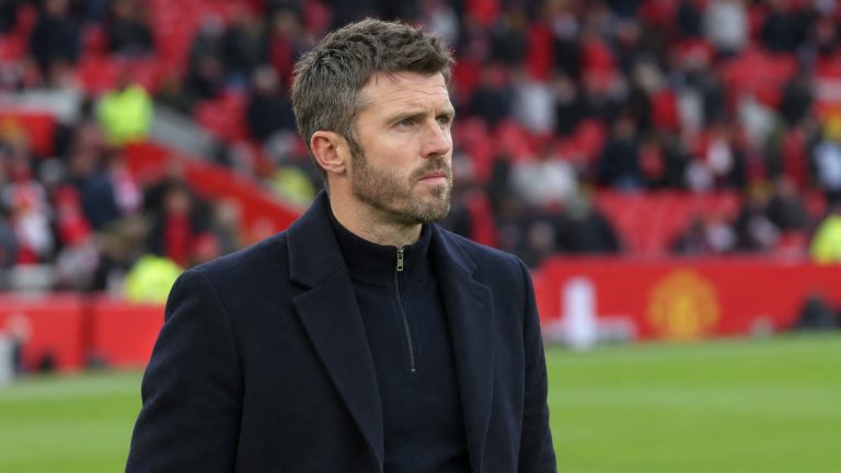 Michael Carrick wearing a dark navy coat beside the pitch with red stadium seating behind him.